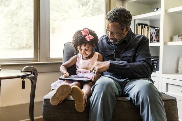 An adult reads with a child seated in front of a window in a home.
